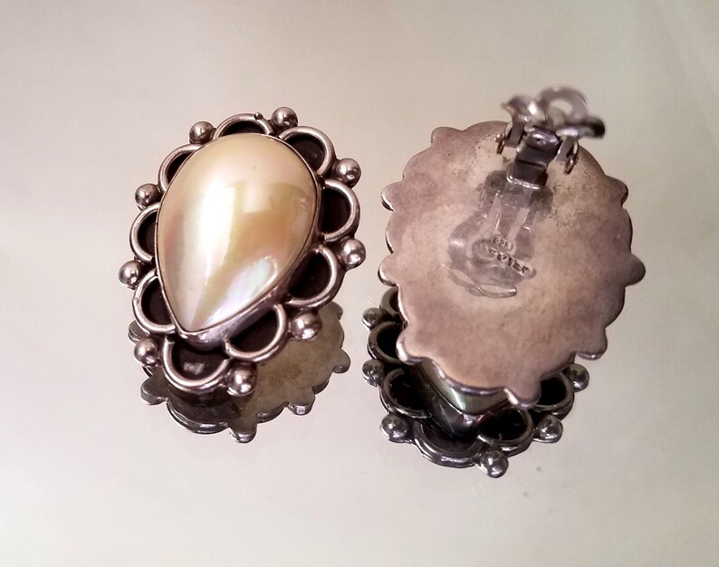 May include: A pair of silver earrings with a pearlescent white teardrop-shaped stone set in a silver filigree design. The earrings have a clip-on closure.