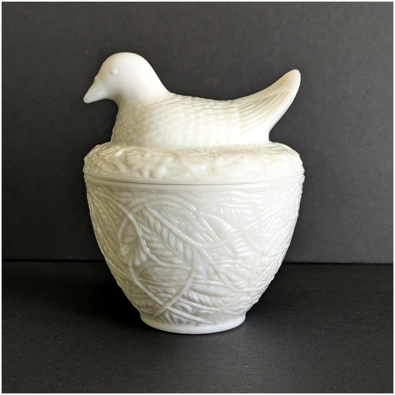 Puede incluir: Una caja de baratijas blanca de estilo vintage con una tapa en forma de p&aacute;jaro. El p&aacute;jaro se asienta sobre una base en forma de nido, y la caja presenta un patr&oacute;n de hojas detallado. Esta pieza decorativa es probablemente de cer&aacute;mica o vidrio.