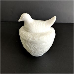 Puede incluir: Una figura de p&aacute;jaro de cer&aacute;mica blanca sobre un recipiente decorativo en forma de huevo. El p&aacute;jaro tiene detalles texturizados en su cuerpo y est&aacute; posado sobre una base en forma de nido. El recipiente presenta un patr&oacute;n de hojas.