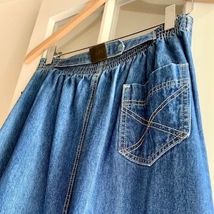 May include: A blue denim skirt with an elastic waistband and a back pocket. The skirt is hanging on a wooden hanger. The pocket has a decorative stitch pattern. The skirt has a belt with a gold-colored buckle.