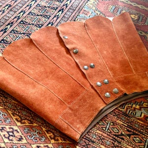 vintage suede skirt in orange rust color with unique scalloped hemline. vintage mini skirt with a series of vertical suede monochrome panels and dark toned brass snaps at front closure. vintage leather skirt. patchwork suede skirt. vintage patchwork skirts. 1960s vintage skirts. vintage 60s skirts. vintage leather patchwork skirt. vintage 1960s mini skirt. vintage suede mini skirts. vintage leather mini skirt. vintage patchwork mini skirt.