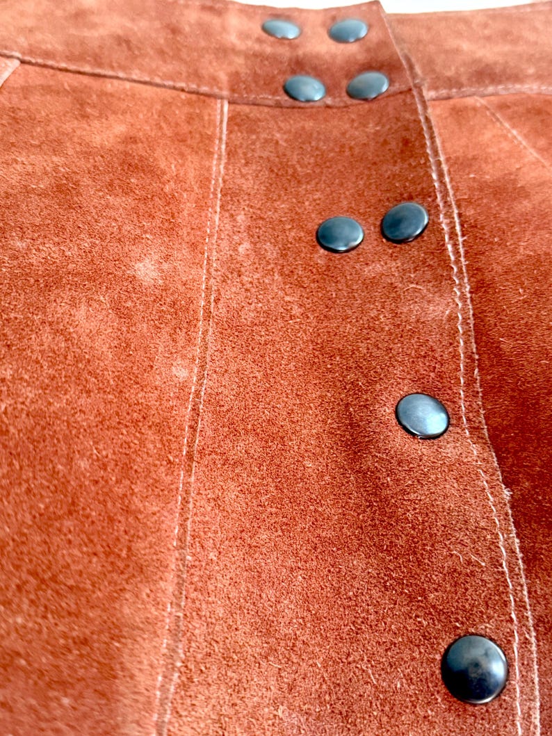 vintage suede skirt in orange rust color with unique scalloped hemline. vintage mini skirt with a series of vertical suede monochrome panels and dark toned brass snaps at front closure. vintage leather skirt. patchwork suede skirt. vintage patchwork skirts. 1960s vintage skirts. vintage 60s skirts. vintage leather patchwork skirt. vintage 1960s mini skirt. vintage suede mini skirts. vintage leather mini skirt. vintage patchwork mini skirt.
