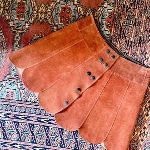 vintage suede skirt in orange rust color with unique scalloped hemline. vintage mini skirt with a series of vertical suede monochrome panels and dark toned brass snaps at front closure. vintage leather skirt. patchwork suede skirt. vintage patchwork skirts. 1960s vintage skirts. vintage 60s skirts. vintage leather patchwork skirt. vintage 1960s mini skirt. vintage suede mini skirts. vintage leather mini skirt. vintage patchwork mini skirt.