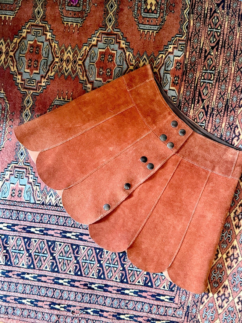 vintage suede skirt in orange rust color with unique scalloped hemline. vintage mini skirt with a series of vertical suede monochrome panels and dark toned brass snaps at front closure. vintage leather skirt. patchwork suede skirt. vintage patchwork skirts. 1960s vintage skirts. vintage 60s skirts. vintage leather patchwork skirt. vintage 1960s mini skirt. vintage suede mini skirts. vintage leather mini skirt. vintage patchwork mini skirt.