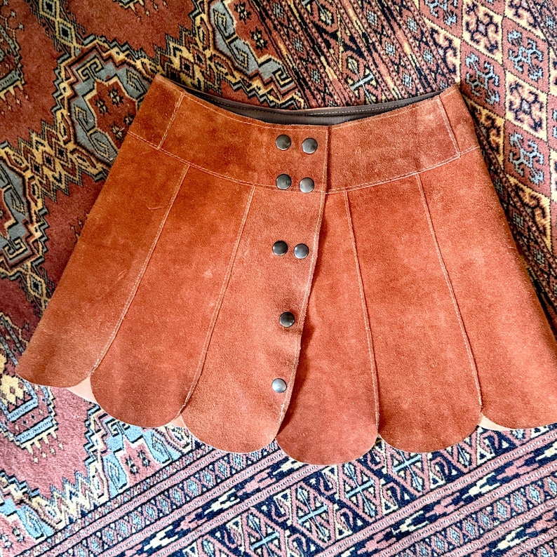 vintage suede skirt in orange rust color with unique scalloped hemline. vintage mini skirt with a series of vertical suede monochrome panels and dark toned brass snaps at front closure. vintage leather skirt. patchwork suede skirt. vintage patchwork skirts. 1960s vintage skirts. vintage 60s skirts. vintage leather patchwork skirt. vintage 1960s mini skirt. vintage suede mini skirts. vintage leather mini skirt. vintage patchwork mini skirt.