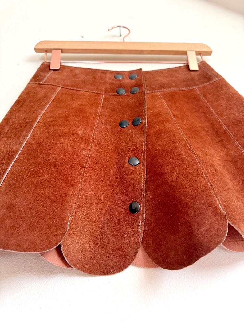 vintage suede skirt in orange rust color with unique scalloped hemline. vintage mini skirt with a series of vertical suede monochrome panels and dark toned brass snaps at front closure. vintage leather skirt. patchwork suede skirt. vintage patchwork skirts. 1960s vintage skirts. vintage 60s skirts. vintage leather patchwork skirt. vintage 1960s mini skirt. vintage suede mini skirts. vintage leather mini skirt. vintage patchwork mini skirt.