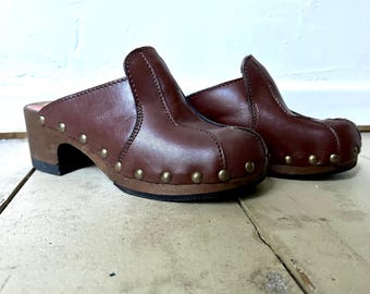 May include: A pair of brown leather clogs with wooden heels and metal studs. The clogs have a rounded toe and a closed back.