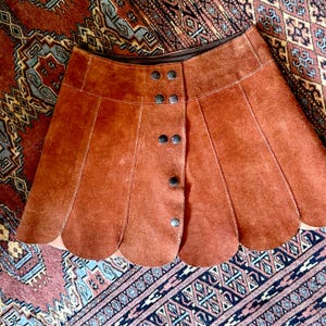 vintage suede skirt in orange rust color with unique scalloped hemline. vintage mini skirt with a series of vertical suede monochrome panels and dark toned brass snaps at front closure. vintage leather skirt. patchwork suede skirt. vintage patchwork skirts. 1960s vintage skirts. vintage 60s skirts. vintage leather patchwork skirt. vintage 1960s mini skirt. vintage suede mini skirts. vintage leather mini skirt. vintage patchwork mini skirt.