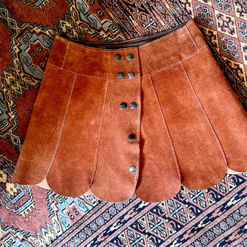 vintage suede skirt in orange rust color with unique scalloped hemline. vintage mini skirt with a series of vertical suede monochrome panels and dark toned brass snaps at front closure. vintage leather skirt. patchwork suede skirt. vintage patchwork skirts. 1960s vintage skirts. vintage 60s skirts. vintage leather patchwork skirt. vintage 1960s mini skirt. vintage suede mini skirts. vintage leather mini skirt. vintage patchwork mini skirt.