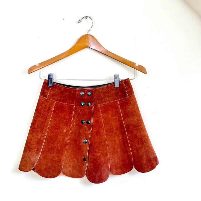 vintage suede skirt in orange rust color with unique scalloped hemline. vintage mini skirt with a series of vertical suede monochrome panels and dark toned brass snaps at front closure. vintage leather skirt. patchwork suede skirt. vintage patchwork skirts. 1960s vintage skirts. vintage 60s skirts. vintage leather patchwork skirt. vintage 1960s mini skirt. vintage suede mini skirts. vintage leather mini skirt. vintage patchwork mini skirt.