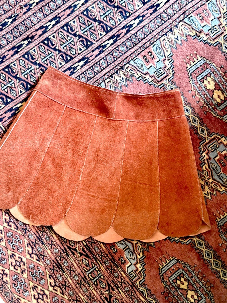 vintage suede skirt in orange rust color with unique scalloped hemline. vintage mini skirt with a series of vertical suede monochrome panels and dark toned brass snaps at front closure. vintage leather skirt. patchwork suede skirt. vintage patchwork skirts. 1960s vintage skirts. vintage 60s skirts. vintage leather patchwork skirt. vintage 1960s mini skirt. vintage suede mini skirts. vintage leather mini skirt. vintage patchwork mini skirt.
