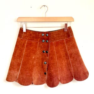 vintage suede skirt in orange rust color with unique scalloped hemline. vintage mini skirt with a series of vertical suede monochrome panels and dark toned brass snaps at front closure. vintage leather skirt. patchwork suede skirt. vintage patchwork skirts. 1960s vintage skirts. vintage 60s skirts. vintage leather patchwork skirt. vintage 1960s mini skirt. vintage suede mini skirts. vintage leather mini skirt. vintage patchwork mini skirt.