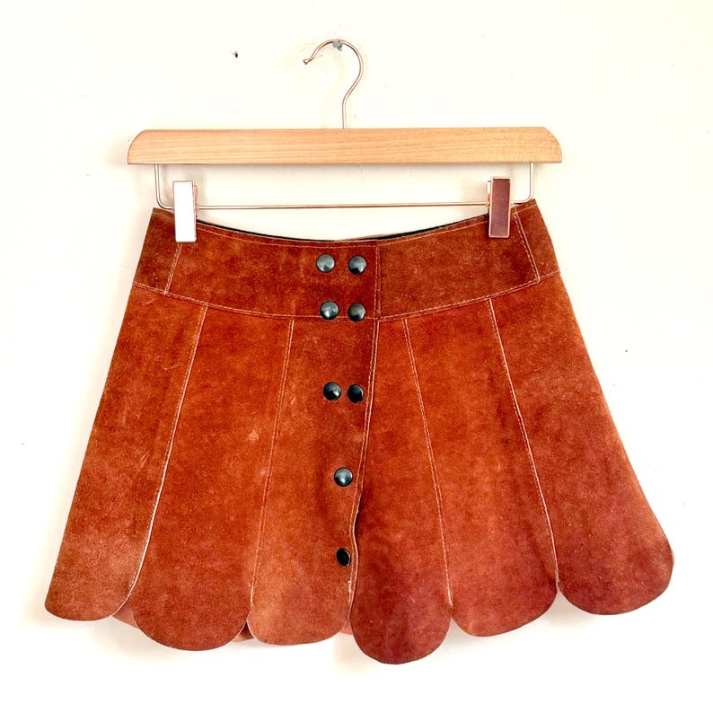vintage suede skirt in orange rust color with unique scalloped hemline. vintage mini skirt with a series of vertical suede monochrome panels and dark toned brass snaps at front closure. vintage leather skirt. patchwork suede skirt. vintage patchwork skirts. 1960s vintage skirts. vintage 60s skirts. vintage leather patchwork skirt. vintage 1960s mini skirt. vintage suede mini skirts. vintage leather mini skirt. vintage patchwork mini skirt.