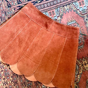vintage suede skirt in orange rust color with unique scalloped hemline. vintage mini skirt with a series of vertical suede monochrome panels and dark toned brass snaps at front closure. vintage leather skirt. patchwork suede skirt. vintage patchwork skirts. 1960s vintage skirts. vintage 60s skirts. vintage leather patchwork skirt. vintage 1960s mini skirt. vintage suede mini skirts. vintage leather mini skirt. vintage patchwork mini skirt.