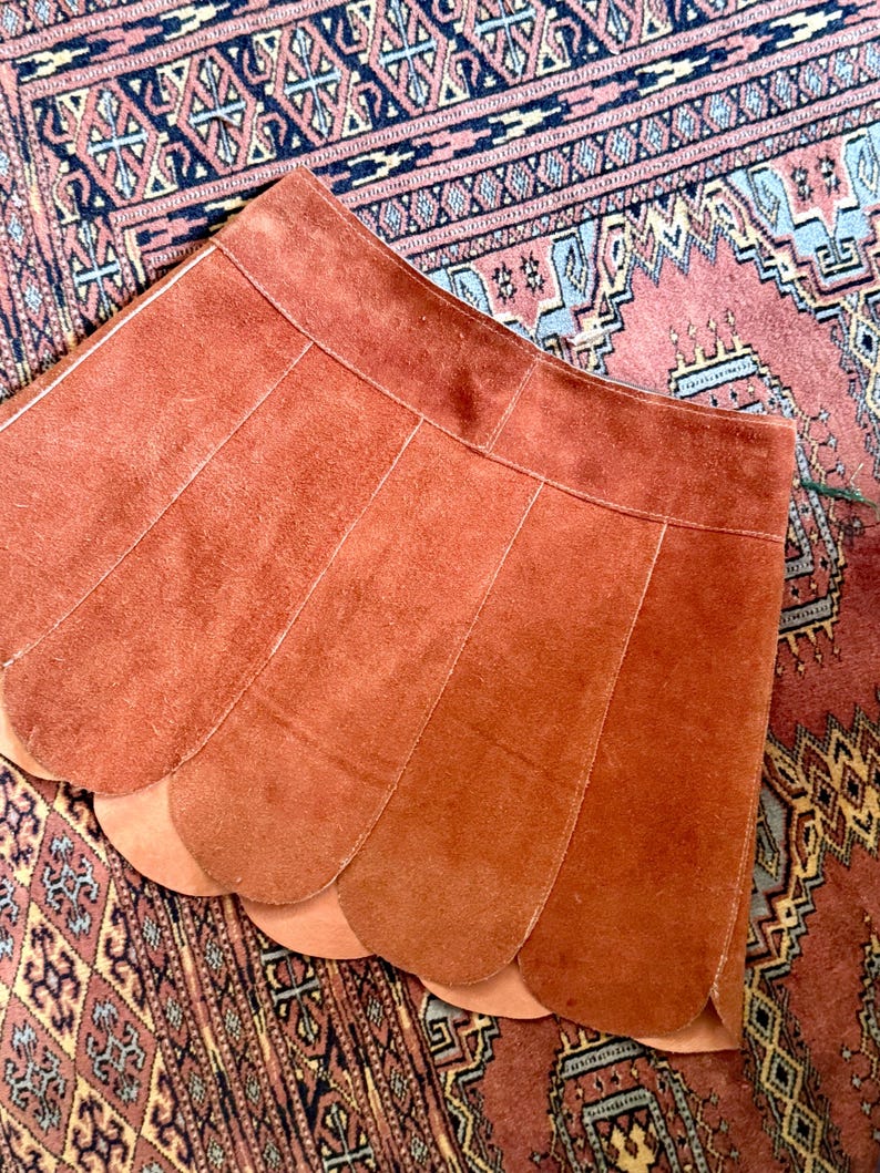 vintage suede skirt in orange rust color with unique scalloped hemline. vintage mini skirt with a series of vertical suede monochrome panels and dark toned brass snaps at front closure. vintage leather skirt. patchwork suede skirt. vintage patchwork skirts. 1960s vintage skirts. vintage 60s skirts. vintage leather patchwork skirt. vintage 1960s mini skirt. vintage suede mini skirts. vintage leather mini skirt. vintage patchwork mini skirt.