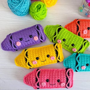 Puede incluir: Seis juguetes de crayón de crochet con caras sonrientes. Los crayones son morado, amarillo, verde, naranja, azul y rosa.