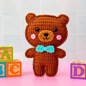 May include: A handmade brown crochet teddy bear with a light brown muzzle, pink cheeks, and a blue bow tie. The bear is standing in front of colorful wooden alphabet blocks. The blocks spell out the letters A, B, C, and D.