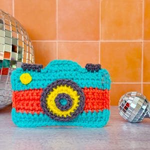 Puede incluir: Un juguete de cámara tejido a ganchillo con un cuerpo de color turquesa brillante y una franja horizontal naranja en el centro. El objetivo está formado por círculos concéntricos en gris oscuro, amarillo y turquesa. Tiene botones grises.