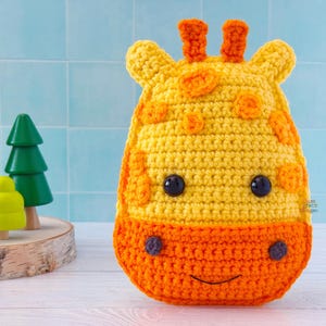 May include: A handmade crochet giraffe toy in yellow and orange. The giraffe has black button eyes, small orange horns, and a smiling mouth. The toy is sitting on a white surface with wooden toy trees in the background.