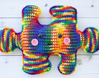 May include: A colorful crocheted puzzle piece toy with a smiling face. The toy is made with rainbow stripes of yarn and has black button eyes and pink cheeks.