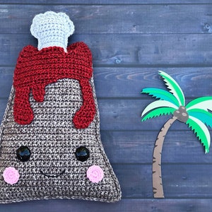 May include: A crocheted volcano toy with a red lava flow, white smoke, and a smiling face. The volcano is next to a paper craft palm tree on a blue wooden background.