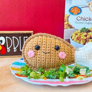 May include: A crocheted turkey leg with a smiling face and pink cheeks sits on a plate with salad. A sign that says "Gobble" is in the background, along with a box of chicken-flavored stuffing mix.