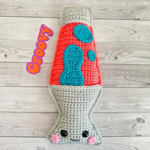May include: A crocheted lava lamp plush toy with a red base, blue lava, and a gray base. The toy has a smiling face with black eyes and pink cheeks. The word "Groovy" is written in pink and orange letters on a purple background.