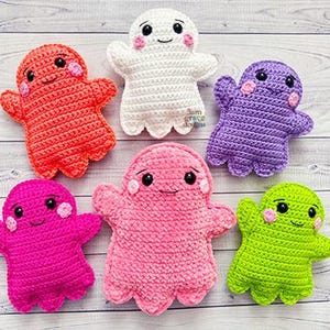 May include: A collection of six handmade crochet ghost dolls in various colors, including red, white, purple, pink, and green. Each ghost has a smiling face with black eyes and pink cheeks. The dolls are arranged on a light wood-grain surface.