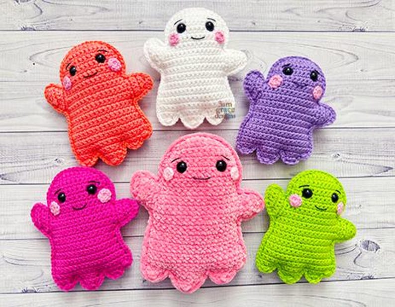 May include: A collection of six handmade crochet ghost dolls in various colors, including red, white, purple, pink, and green. Each ghost has a smiling face with black eyes and pink cheeks. The dolls are arranged on a light wood-grain surface.
