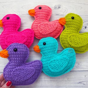 Peut inclure: Cinq canards en crochet de diff&eacute;rentes couleurs&nbsp;: rose, violet, vert, bleu et turquoise. Les canards sont dispos&eacute;s en rang&eacute;e sur une surface en bois blanc.