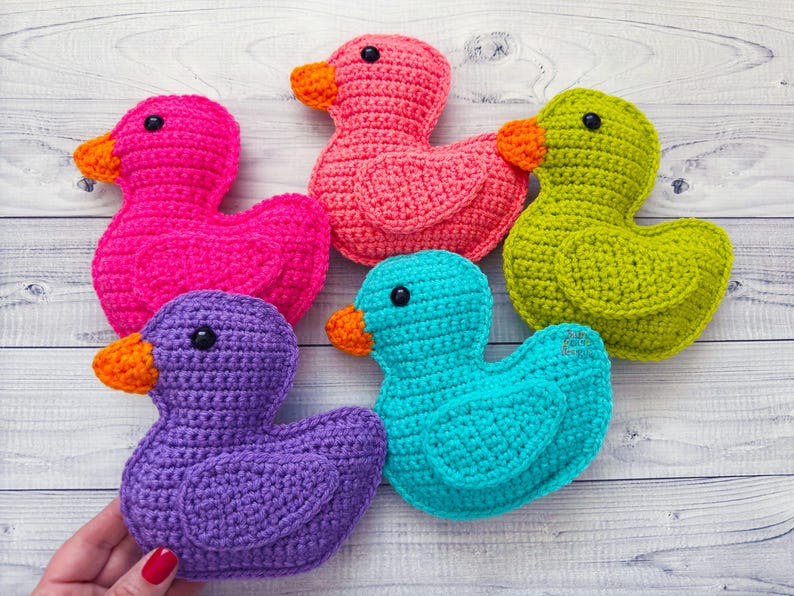 Peut inclure: Cinq canards en crochet de diff&eacute;rentes couleurs&nbsp;: rose, violet, vert, bleu et turquoise. Les canards sont dispos&eacute;s en rang&eacute;e sur une surface en bois blanc.
