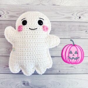 May include: A handmade white crochet ghost with black eyes, a smiling mouth, and pink cheeks. A pink and gray pumpkin decoration is next to the ghost. The items are on a light wood grain surface. Halloween themed.