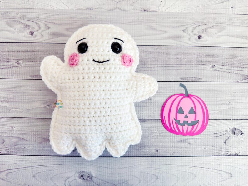 May include: A handmade white crochet ghost with black eyes, a smiling mouth, and pink cheeks. A pink and gray pumpkin decoration is next to the ghost. The items are on a light wood grain surface. Halloween themed.
