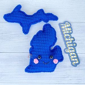 Op de afbeelding: Twee blauwe gehaakte Michigan-staatsvormen, één met een lachend gezicht en roze wangen. Het woord "Michigan" staat op een blauw en geel bord. De items liggen op een wit houtnerfoppervlak.