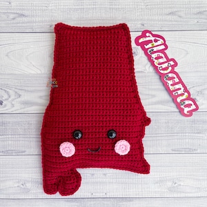 May include: Red crocheted state of Alabama pillow with a smiling face, black eyes, and pink cheeks. The pillow has the word "Alabama" written on a pink tag.