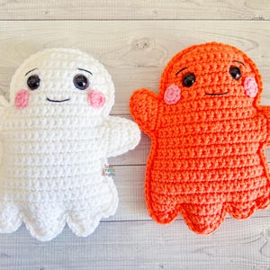 May include: Two handmade crochet ghost plushies. One is white, the other is orange. Both have black eyes, pink cheeks, and smiling mouths. The ghost plushies have outstretched arms and a scalloped bottom edge.