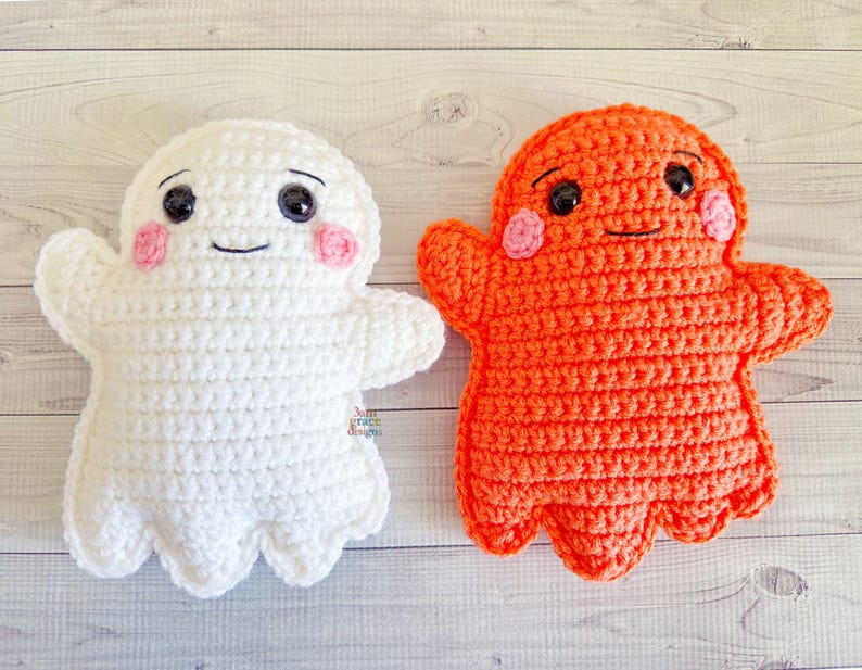 May include: Two handmade crochet ghost plushies. One is white, the other is orange. Both have black eyes, pink cheeks, and smiling mouths. The ghost plushies have outstretched arms and a scalloped bottom edge.