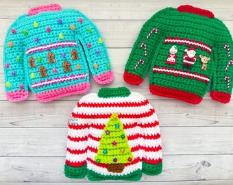 May include: Three crocheted Christmas sweaters. One is blue with gingerbread men, one is green with candy canes and a Santa Claus, and one is red and white striped with a green Christmas tree.