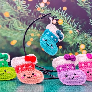 May include: A collection of colorful, crocheted Christmas stocking ornaments. The stockings are in shades of green, pink, purple, and blue, each with a white cuff and a bow. They have smiling faces and are decorated with stars.