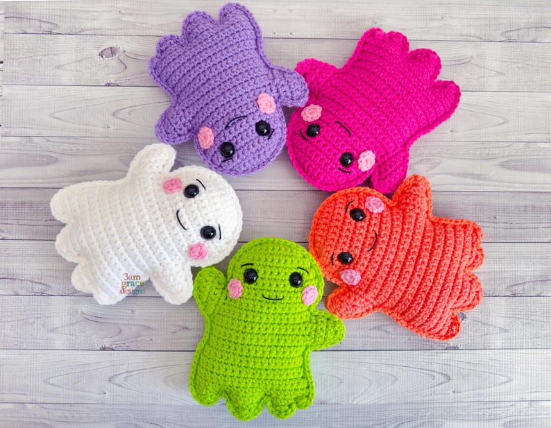 May include: Five colorful, handmade crochet plush toys in the shape of friendly ghosts and monsters. The toys are in shades of white, purple, pink, orange, and green. Each has black eyes and pink cheeks, with a smiling mouth.