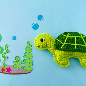 Puede incluir: Una tortuga de ganchillo hecha a mano en tonos verdes y amarillos, con un patrón de caparazón detallado. Sobre un fondo azul claro, algas y burbujas recortadas en papel crean una escena submarina.