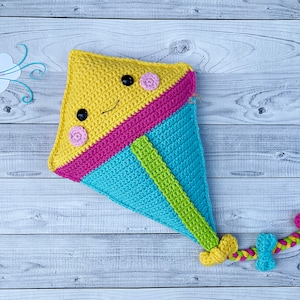 Puede incluir: Un juguete de cometa de crochet en amarillo, rosa, azul y verde. La cometa tiene una cara sonriente con ojos negros y mejillas rosas. La cometa tiene una cola con un lazo rosa e hilo amarillo y verde.
