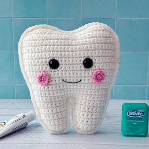 Peut inclure: Une grande dent blanche crochetée avec des yeux en boutons noirs, des joues roses crochetées et une bouche souriante. Un tube de dentifrice et un contenant de fil dentaire sont à côté de la dent. Le fond est un carrelage bleu clair.