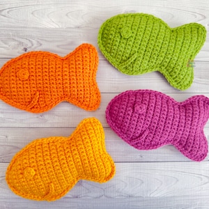 May include: Four crocheted fish toys in different colors: orange, yellow, green, and pink. The fish are lying on a white wooden surface.