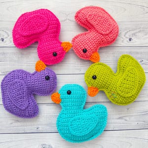 Peut inclure: Quatre canards en peluche crochet&eacute;s de diff&eacute;rentes couleurs&nbsp;: rose, violet, vert et bleu. Les canards sont dispos&eacute;s en cercle sur un fond en bois blanc.
