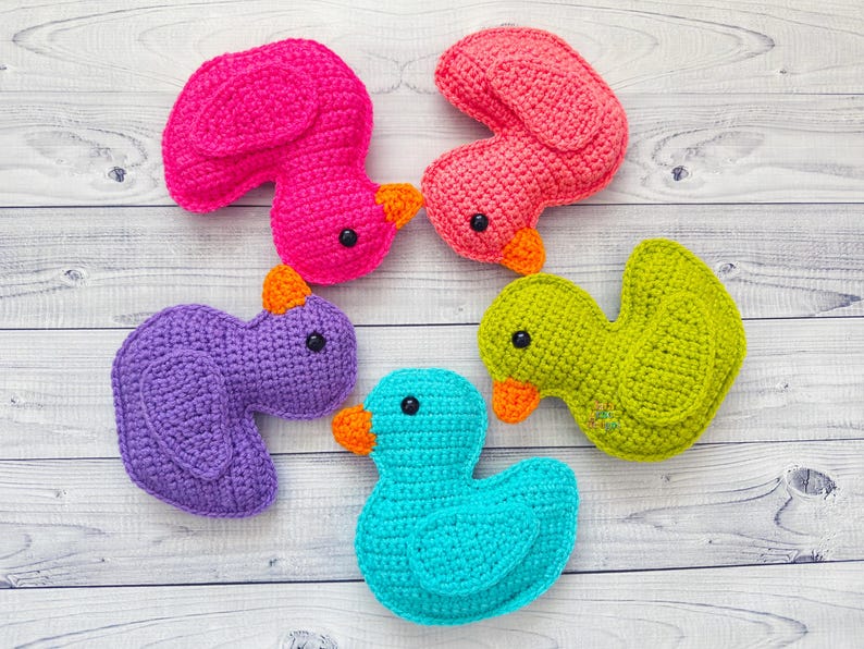 Peut inclure: Quatre canards en peluche crochet&eacute;s de diff&eacute;rentes couleurs&nbsp;: rose, violet, vert et bleu. Les canards sont dispos&eacute;s en cercle sur un fond en bois blanc.
