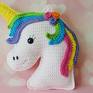 Puede incluir: Una cabeza de unicornio de crochet blanco con una melena arcoíris y tres flores de crochet rosas y naranjas. El unicornio tiene ojos negros y una nariz negra.