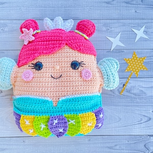 Puede incluir: Un juguete de peluche tejido a crochet con forma de hada con cabello rosado, un vestido azul y una corona. El hada tiene ojos grandes y una cara sonriente. El juguete está sentado sobre una superficie de madera.