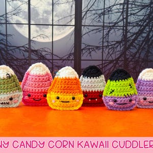 May include: Six crocheted Halloween-themed characters, each resembling candy corn, with smiling faces. The characters are in various colours: green, pink, orange, black, red, and purple, with white tops. They are displayed on an orange surface.