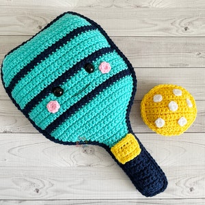 May include: A handmade crochet pickleball set featuring a teal and navy blue paddle with a smiling face and a yellow ball with white dots. The paddle is approximately 12 inches long.
